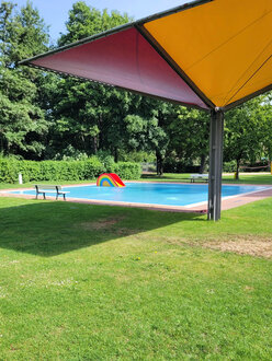 Kleinkindbecken im Freibad Garmissen ohne Personen. Am Beckenrand steht eine Bank, im Becken eine kleine Rutsche in Regenbogenoptik. 
Im Vordergrund ist ein gro�er Regensammelschirm von der Expo 2000 in Hannover zu sehen, der jetzt in Garmissen Schatten spendet.